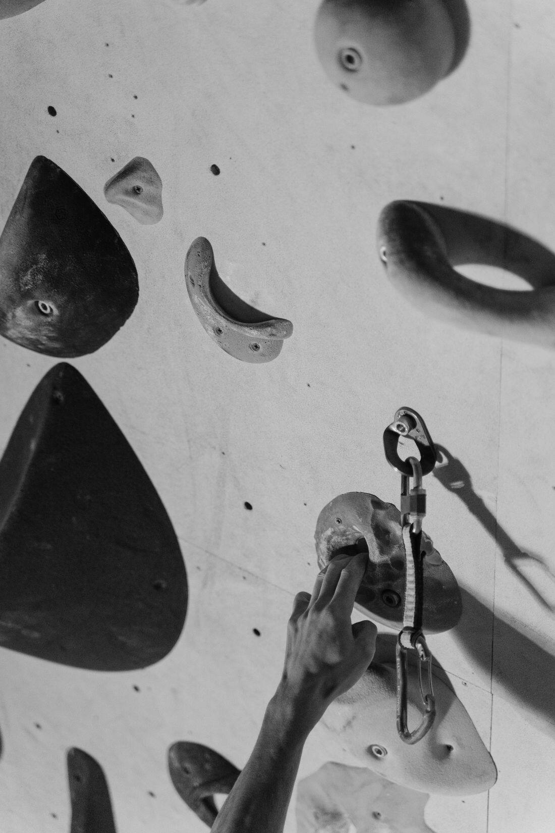 A chalked hand reaching for a hold on an indoor climbing wall beside a clipped quickdraw