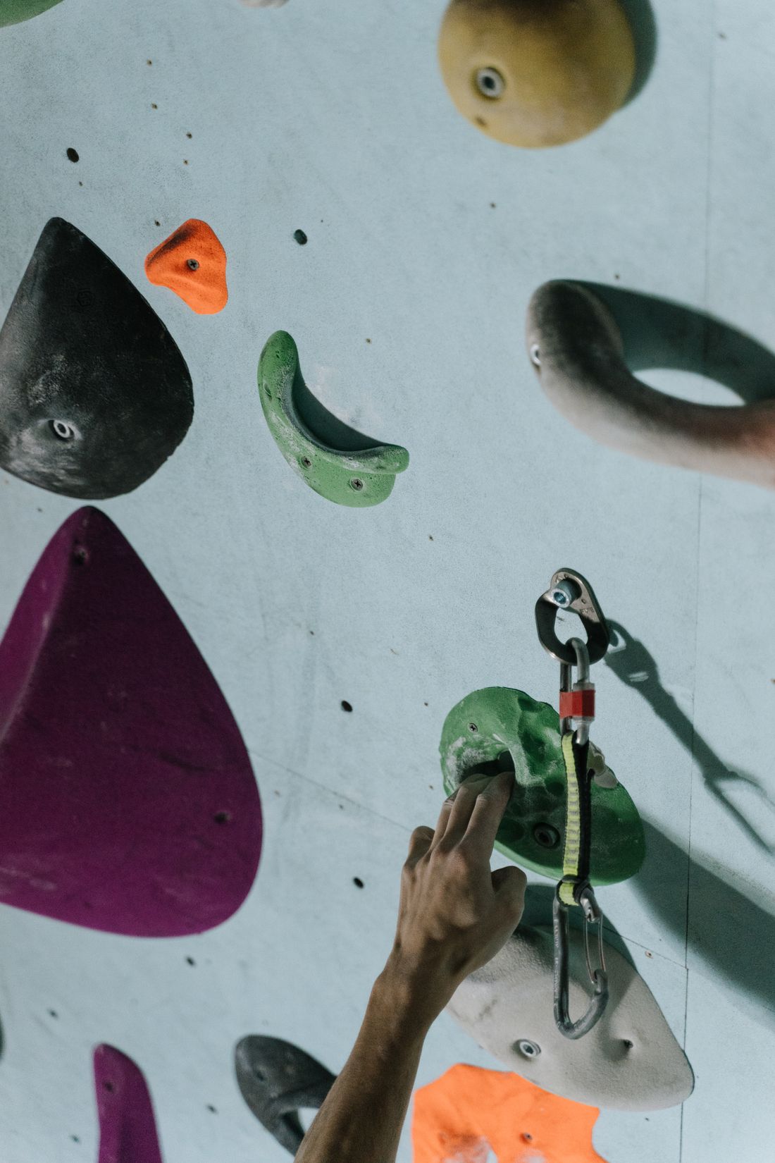 A chalked hand reaching for a hold on an indoor climbing wall beside a clipped quickdraw